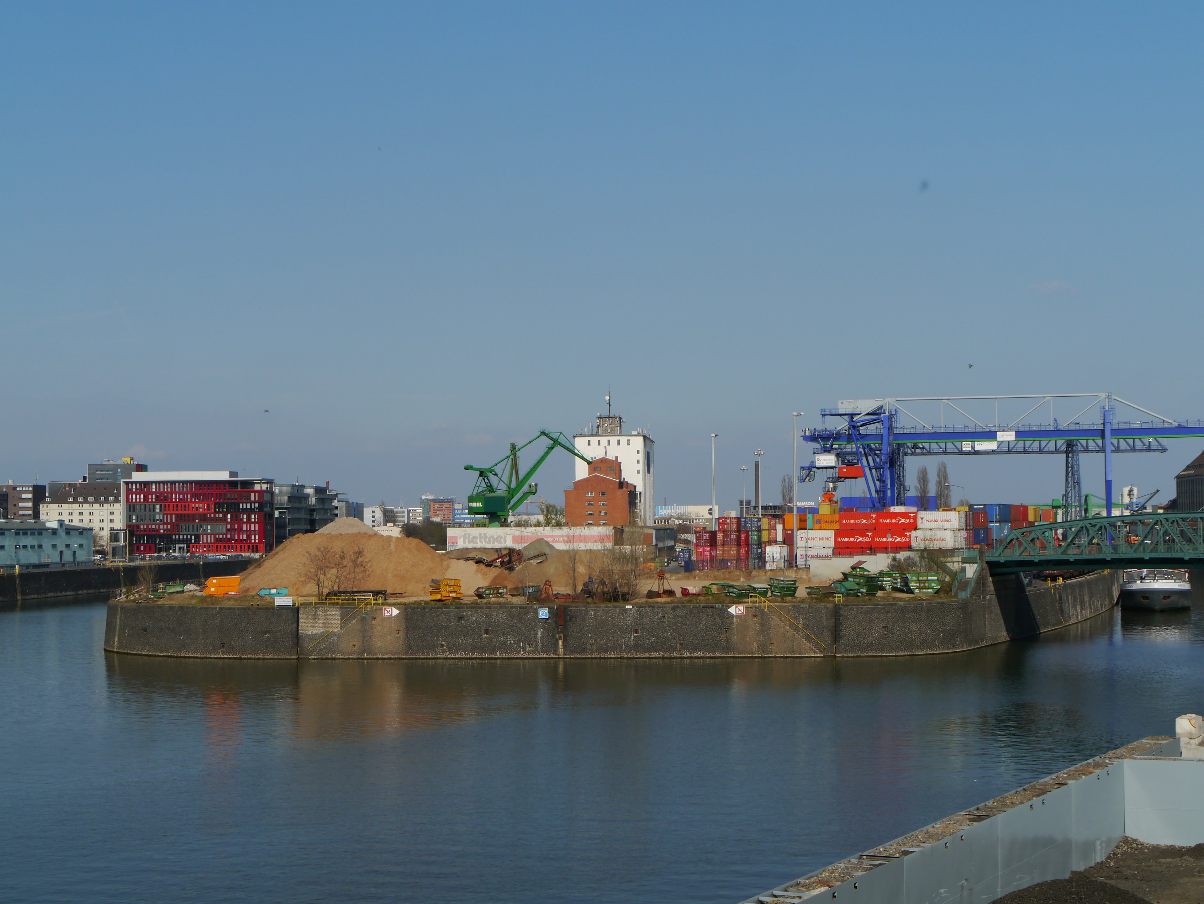 Eine Hafenszene mit einem großen Kran, einem Schiff und verschiedenen Containern, alles vor einem klaren blauen Himmel.